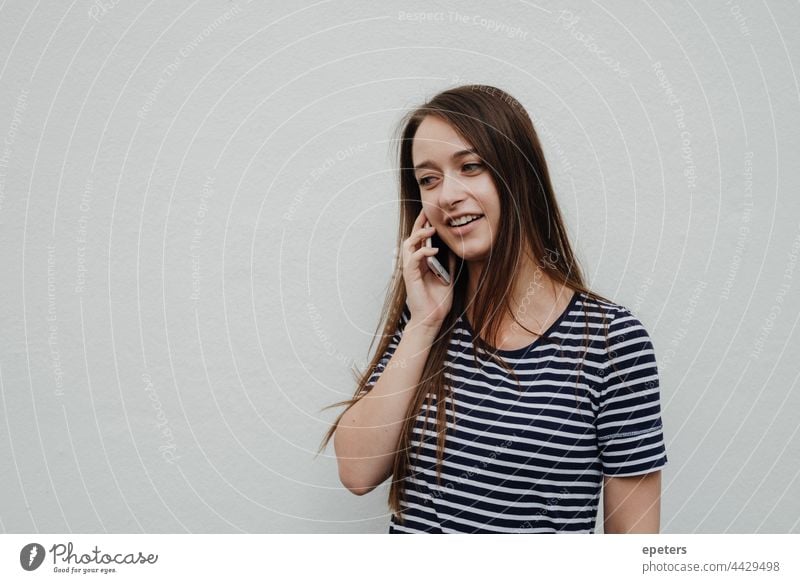 Young woman holding a smartphone to the ear and standing in front of a white wall brown hair casual clothes copy space gen z getting news millenial