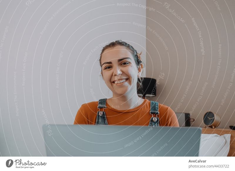 Young woman using his laptop while on bed while smiling, working from home, hipster style, minimal white house computer technology bedroom relaxation portrait