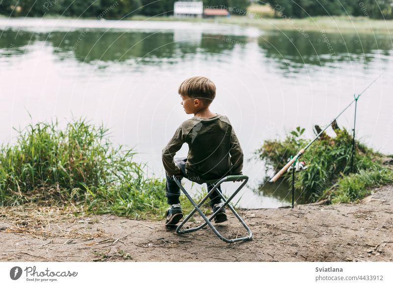 The boy is sitting on a folding chair on the shore of a lake or river. Recreation, weekends, tourism. Rear view child summer kid recreation lifestyle leisure