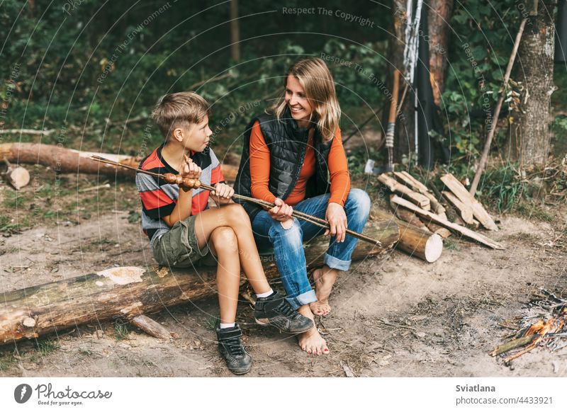 A mother and her teenage son are sitting on a tree in campsites and trying grilled sausages cooked on a fire camping family mom man spending time together