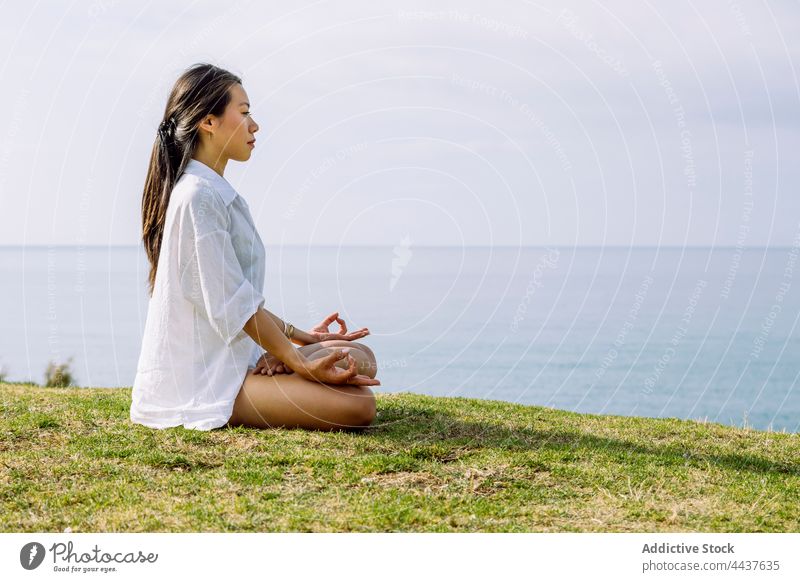 Mindful Asian woman meditating in Lotus pose on green shore lotus pose meditation yoga zen stress relief spirit mindfulness energy seashore eyes closed vitality