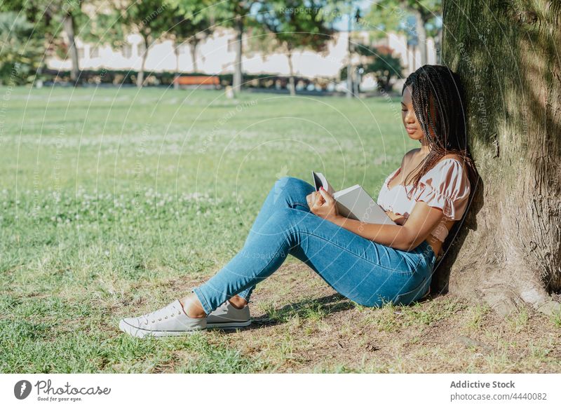 Contemplative ethnic woman with book resting in park contemplate wistful reflective trendy millennial lawn gumshoe crop top textbook literature trunk meadow