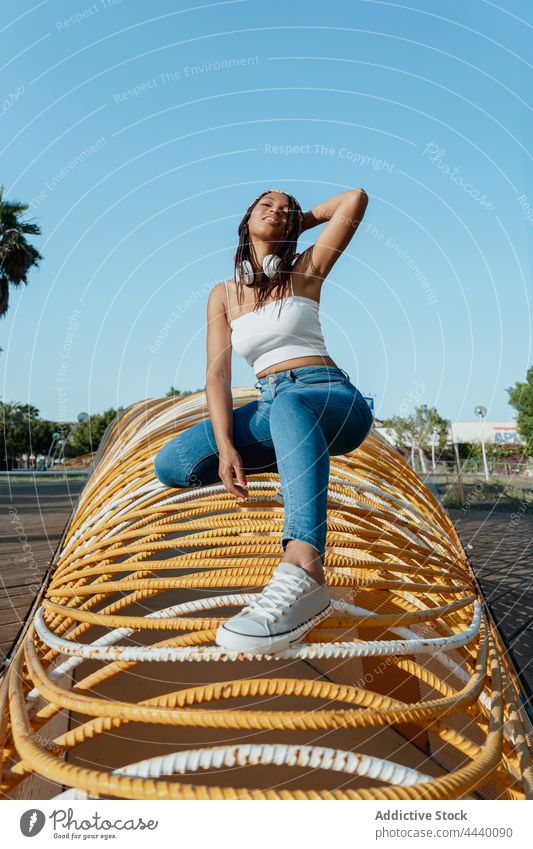 Smiling ethnic woman squatting on geometric construction in town trendy cool individuality cheerful enjoy architecture geometry street millennial headset