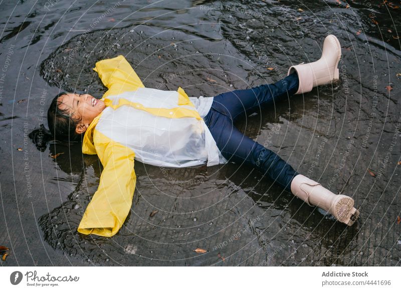 Happy ethnic girl in raincoat having fun in puddle carefree laugh legs apart arms apart childhood pastime rainy weekend spare time reflection pavement gumboots