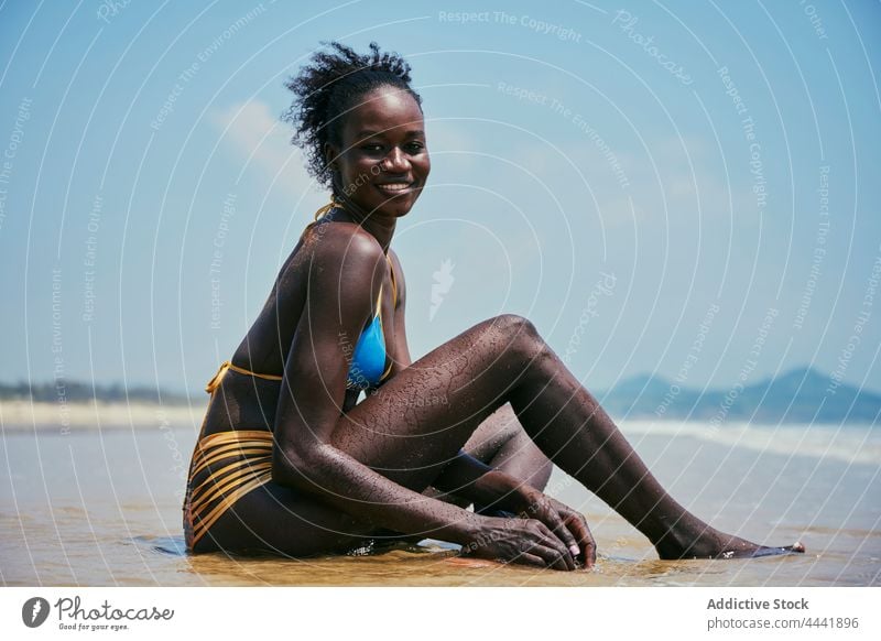 Sensual black model in swimsuit resting on sea shore swimwear body sensual cheerful feminine beauty seashore blue sky woman afro cloudy content lean on hand