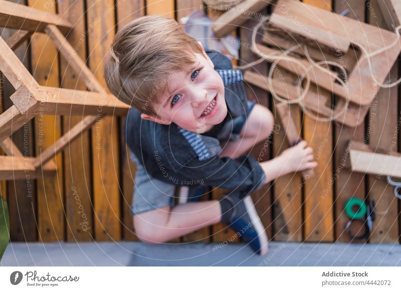 Little boy on boardwalk with wooden pieces childhood gaze legs crossed handmade charming portrait block twine stare blue eyes diy handwork woodwork spare time