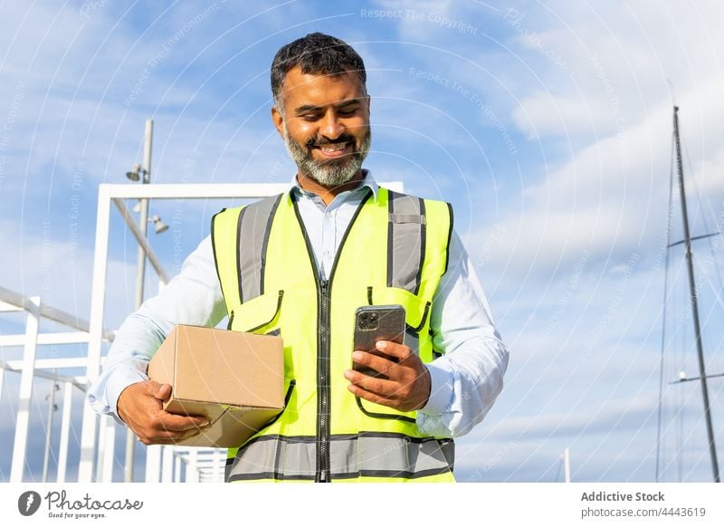 Smiling ethnic workman with box browsing smartphone worker smile happy using carry parcel glad delivery uniform delight watch male occupation job device gadget