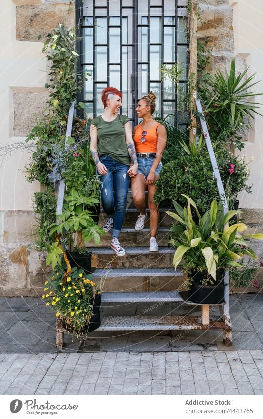 Smiling lesbian couple talking and holding hands standing on stairs speak relationship content spend time cool lgbt women rest tattoo smile interact cheerful