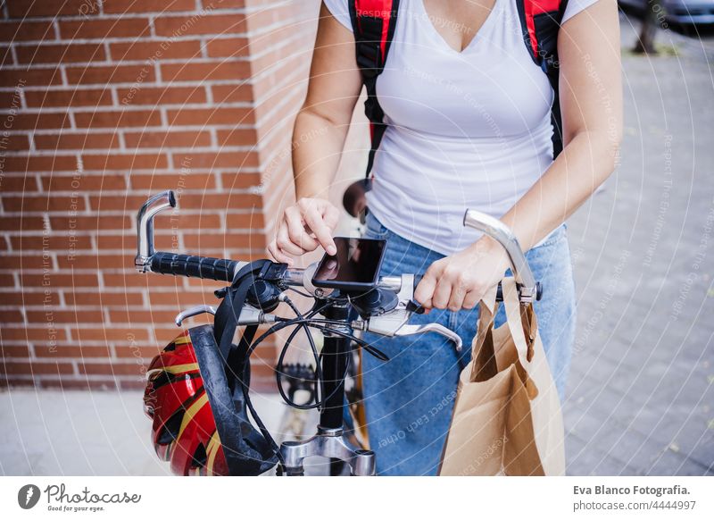 close up of young rider woman wearing red backpack holding paperboard box of food in city. Delivery service concept. Woman checking order in mobile phone