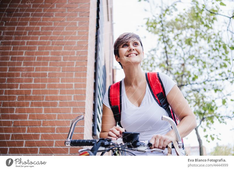 happy young rider woman wearing red backpack holding paperboard box of food in city. Delivery service concept. Woman checking order in mobile phone deliver