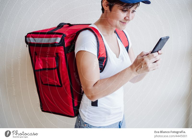 rider woman wearing red backpack delivering food, checking order with smart phone while standing on street in city. Delivery service concept. Sustainable transport