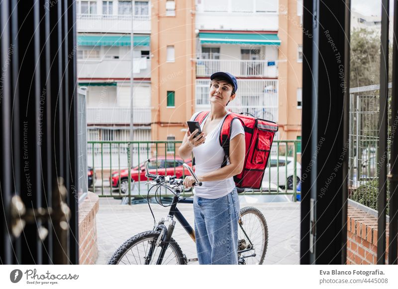 Young rider woman wearing red backpack delivering food on a bike, checking order with smart phone while standing on street gate in city. Delivery service concept. Sustainable transport