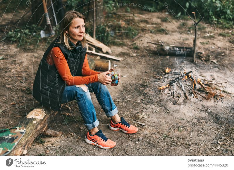 A young woman is warming herself by the fire with a cup of warming tea in the forest drink sitting nature tourist adventure vacation campfire bonfire travel