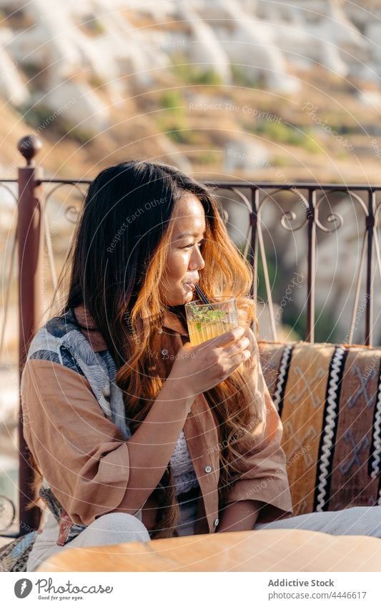 Asian woman drinking mojito on terrace of bar sip straw cocktail glass admire refreshment female beverage balcony sit veranda tasty thirst taste exotic alcohol