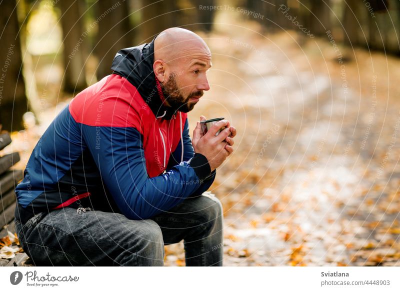 A handsome bearded man is sitting on a bench in an autumn park and drinking hot tea cup coffee fall traveler outdoor casual nature person male season people