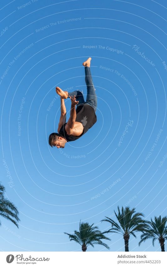 Energetic dancer performing trick in mid air above palm trees backbend jump extreme energy courage adrenalin blue sky man dynamic fearless nature touch leg