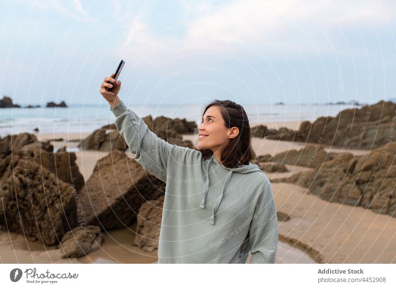 Smiling woman taking selfie on smartphone on rocky beach self portrait seacoast memory moment sky free time using gadget device cellphone watching pastime