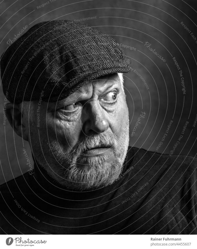 An older man with a beard and cap looks skeptically back into the light. Man portrait Black & white photo Face Looking Masculine Facial hair Light Cap Observe