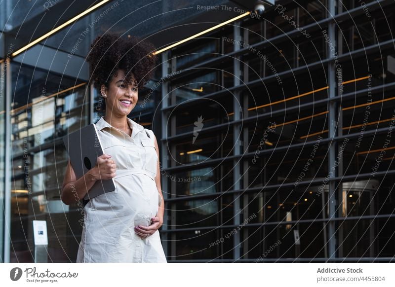 Smiling black woman with laptop coming out of office building pregnant carry maternal anticipate expect tower happy female tummy gadget device belly digital