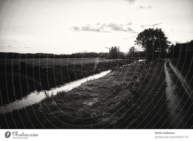 beautiful black and white abstract Latvia landscape. Dirt sand road, water flows in straight river with clean banks, some trees in side atmospheric mood