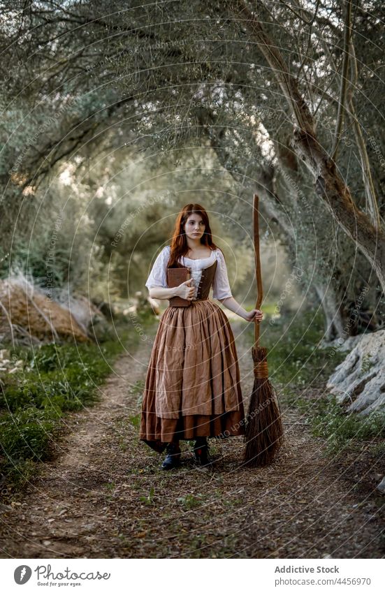 Witch with broomstick and book in forest witch magic woman dress enchantress autumn mystic female witchcraft magician woods stand dull gloomy halloween serious