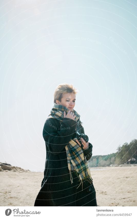 Young woman in a coat and scarft with camera standing on a beach in cold weather, adult autumn blonde caucasian fair weather female glasses happy landscape