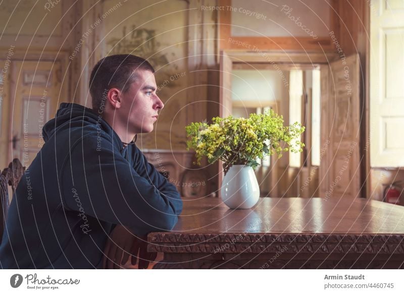 young man in profile sitting at a table with a vase full of yellow flowers portrait room inside interior door old ancient grungy elbows wood serious looking