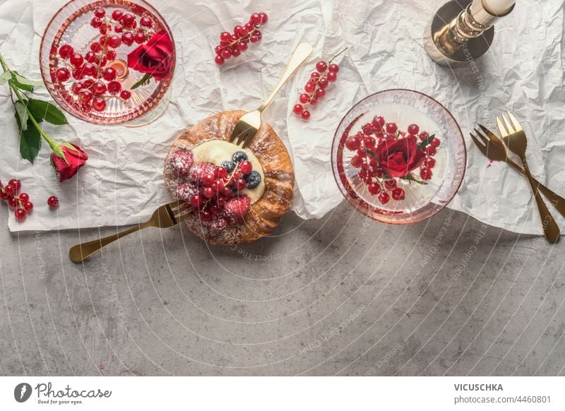 Romantic background with two glasses champagne with red berries and sweet pudding bun, with two gold forks, berries and flowers. Top view rose romantic top view