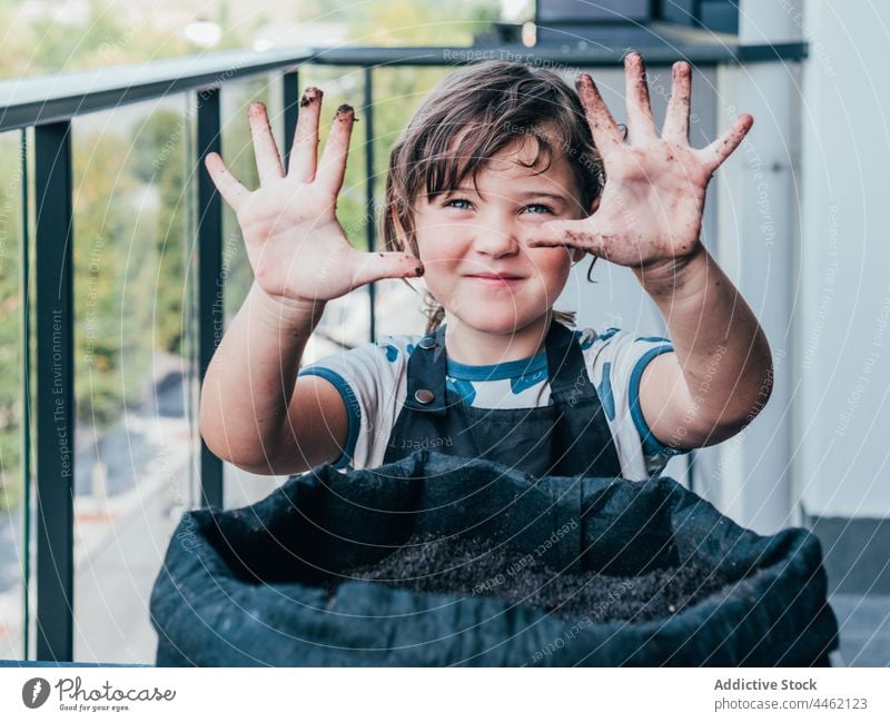 Girl showing dirty hands and looking at camera girl apron pot balcony flowerpot soil kid positive child little flora horticulture demonstrate cheerful daytime