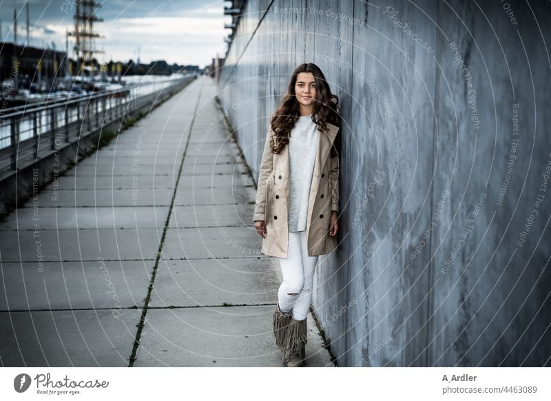 young woman in coat leans against wall, harbour in background youthful Woman Girl Young woman Coat Evening dusk Human being Wall (barrier) Wall (building)