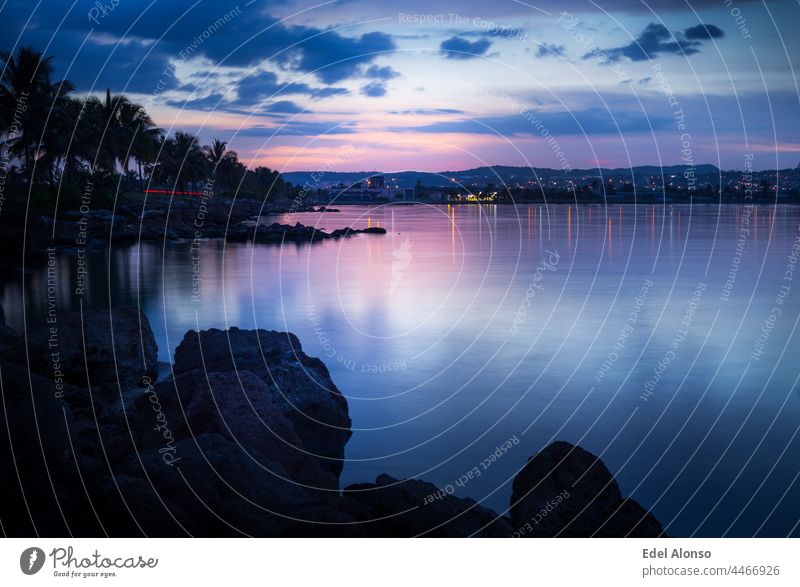 Blue hour in the sunset on the beach in the summertime surrounded by rocks, trees, palms, some mountains in the horizon and a calm water reflecting clouds and lights.