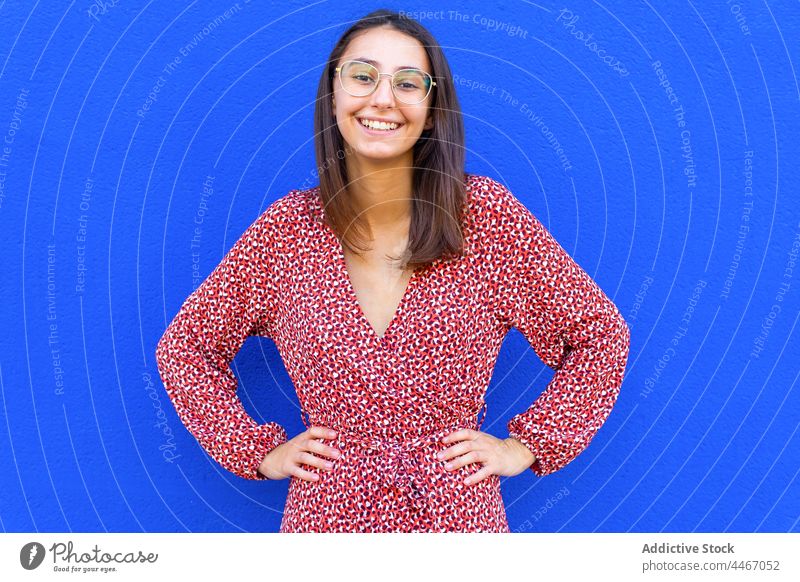 Smiling female standing against blue wall in daytime woman style street dress trendy outfit sidewalk pavement walkway hands on waist casual feminine