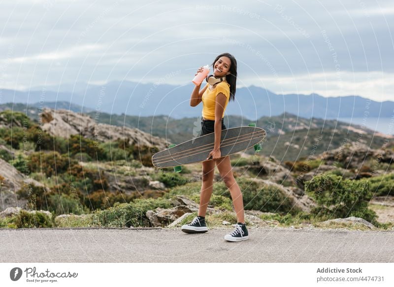 Smiling skater with longboard walking on pavement against mount smile mountain nature sea sport woman skateboarder ethnic latin summer highland ocean ridge