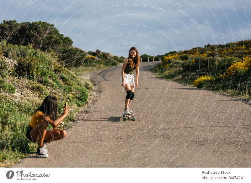 Ethnic woman taking photo of friend balancing on longboard women take photo ride using smartphone hobby activity capture female leisure girlfriend moment grassy