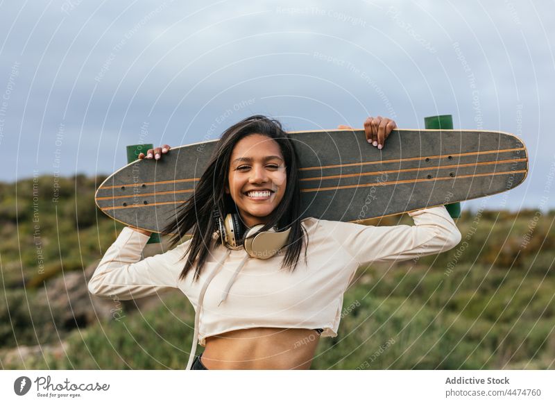 Joyful ethnic woman standing with longboard behind head laugh headphones cool fun playful joy pleasure hobby female smile positive carefree expressive optimist