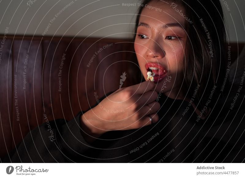 Young Asian woman sitting on sofa and eating popcorn couch at home relax enjoy ethnic calm leisure apartment living room free time positive casual weekend