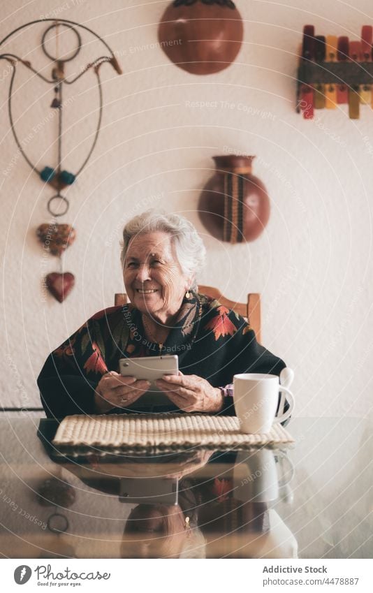 Aged woman with gadget sitting at table tablet at home tea device pensioner senior elderly aged drink browsing using female cup smile warm toothy smile retire