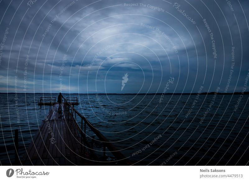 wooden footbridge on lake in sunset blue hour, human silhouette. Trail is made of timber boards, safety railing on right side. Beautiful cloudy sky in soft light