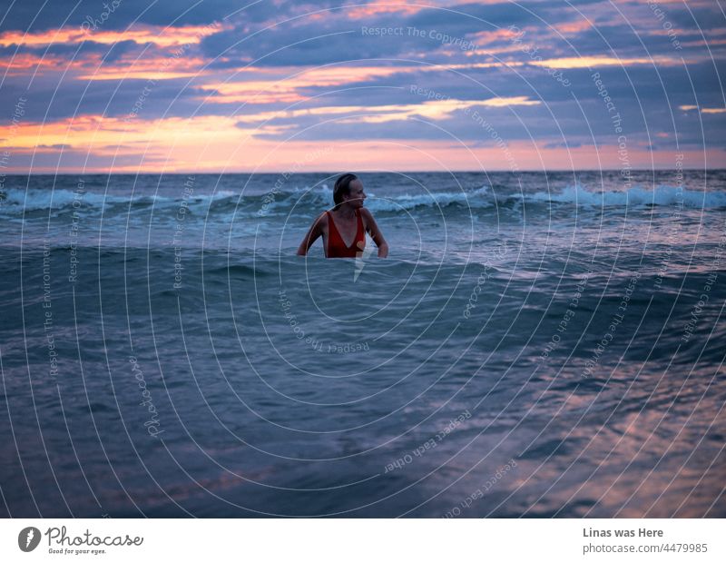 What a gorgeous red-pink-purple sky on a perfect evening by the sea. A pretty woman with a red swimsuit is enjoying the waves. Tempting blue water everywhere you look around. A female model in the sea is alone but doesn’t feel lonely.