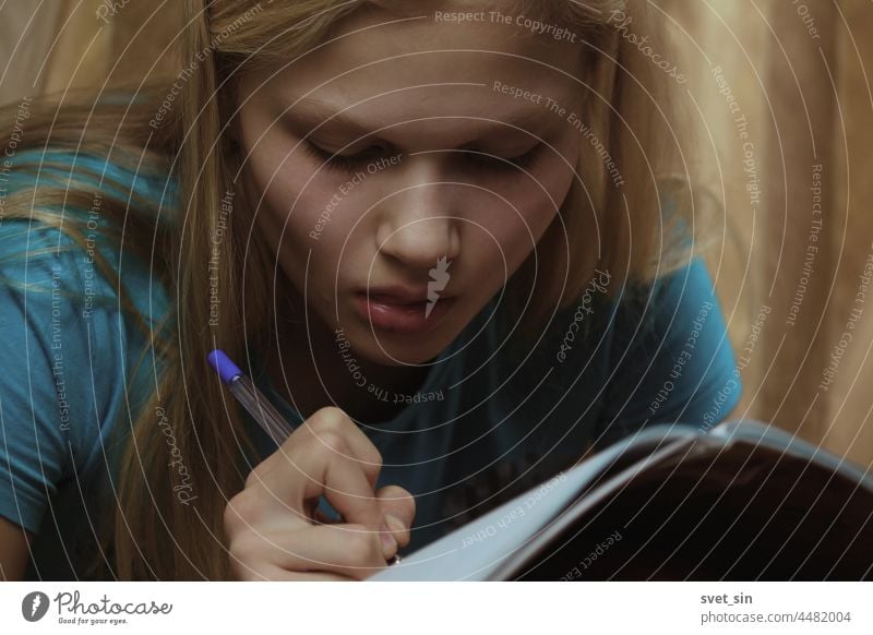 A blonde teenage girl is busy writing in a notebook indoors. write pen concentratedly studying home person caucasian lifestyle homework portrait people sitting