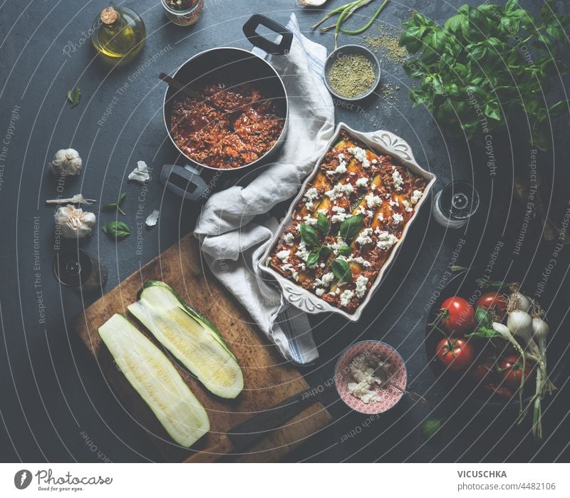 Homemade courgette lasagne with fresh basil, bolognese, tomatoes, garlic, olive oil and cheese in white baking dish on dark concrete kitchen table. Cooking italian food at home. Top view.