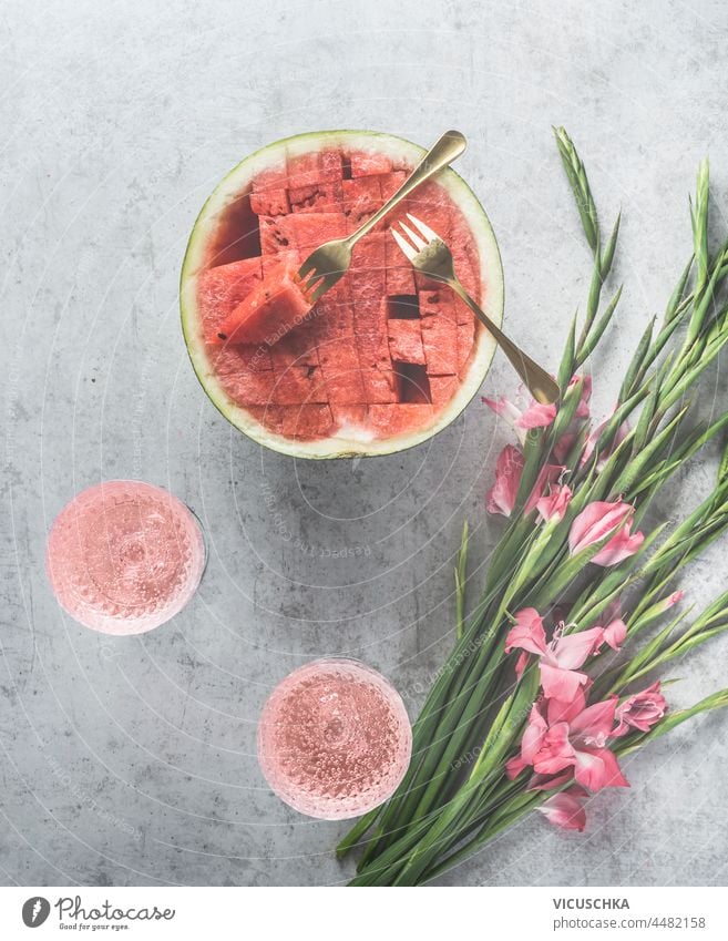 Watermelon cocktails with sparkling wine in wine glasses, sliced watermelon and pink flowers on pale grey concrete kitchen table. Summer celebration with homemade drinks and fruit. Top view.