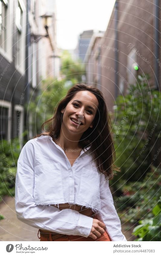 Stylish woman walking on street narrow building plant urban smile trendy exterior pavement female city town stroll confident flora summer path young calm