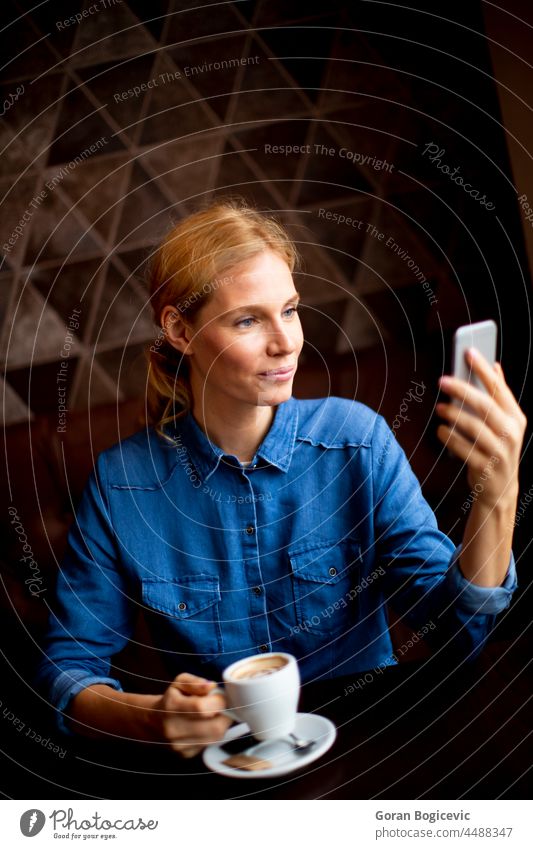 Pretty young woman sitting at a table with coffee or cappuccino and using mobile phone connection communication female technology caucasian smile bar telephone