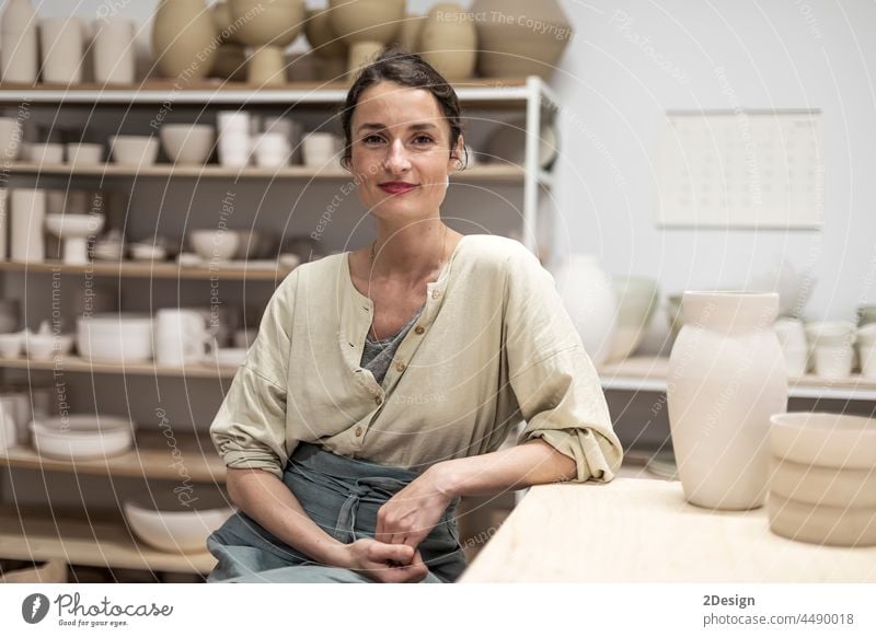 Portrait of beautiful happy craft woman wearing apron and smiling while sitting in her art studio or craft pottery shop workshop ceramist occupation female