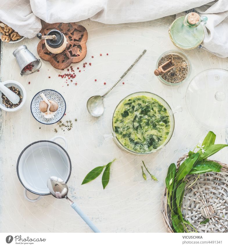 Wild garlic soup on white kitchen table with bowls, ingredients, herbs, spices and kitchen utensils. Cooking with seasonal wild herbs at home. Top view.