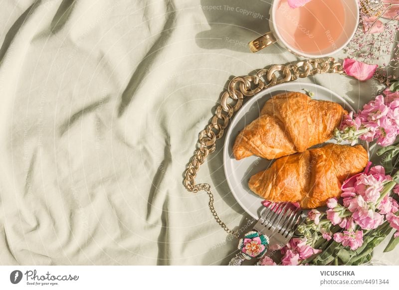 Croissant breakfast background on pale tablecloth with pink tea in tea cup, gold chain and pink flowers. Romantic french sweet breakfast at home. Top view with copy space.