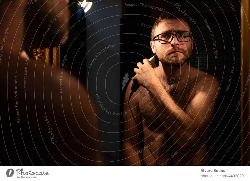 A young man shaving and trimming his beard with an electric razor, in the bathroom at home, early in the morning, to be groomed before going out