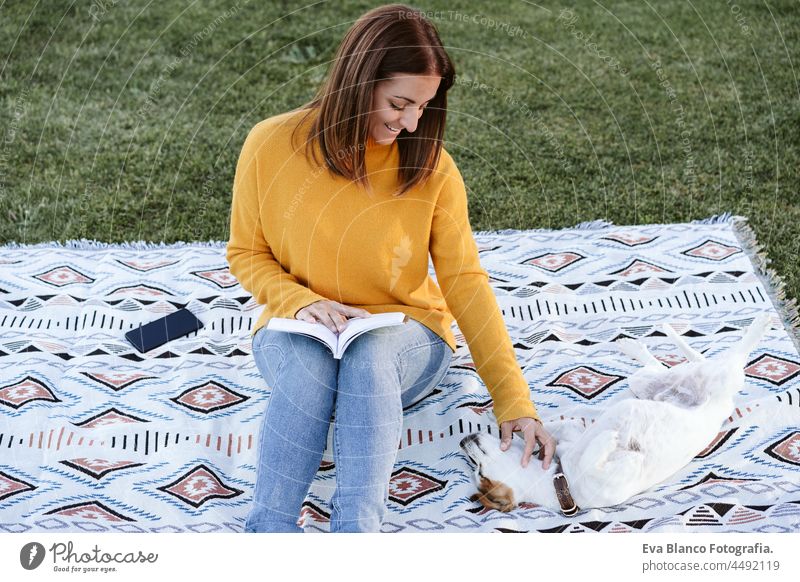 woman having fun with jack russell dog in park, sitting on blanket during autumn season. Woman reading a book while cuddling dog. Pets and love concept relax