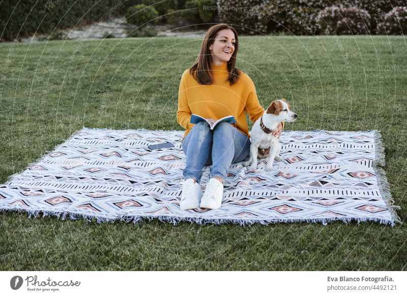 woman having fun with jack russell dog in park, sitting on blanket during autumn season. Woman reading a book while hugging dog. Pets and love concept relax